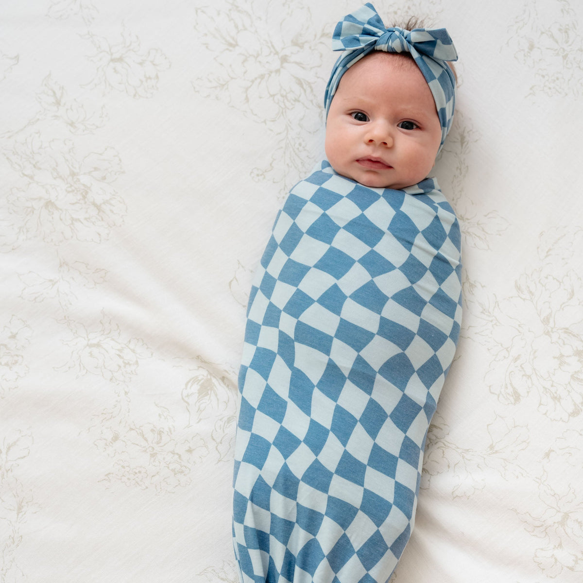 A baby lying on a white surface is swaddled snugly in a blue and white checkered knit blanket, wearing a matching headband with a top knot.