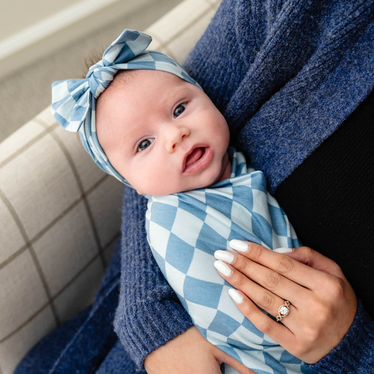 An adult holds a baby swaddled in a blue and white checkered knit blanket, with the baby wearing a matching top-knot headband.