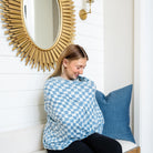 Woman sitting on a couch wrapped in a blue and white checkered blanket, with a decorative mirror and pillow in the background.