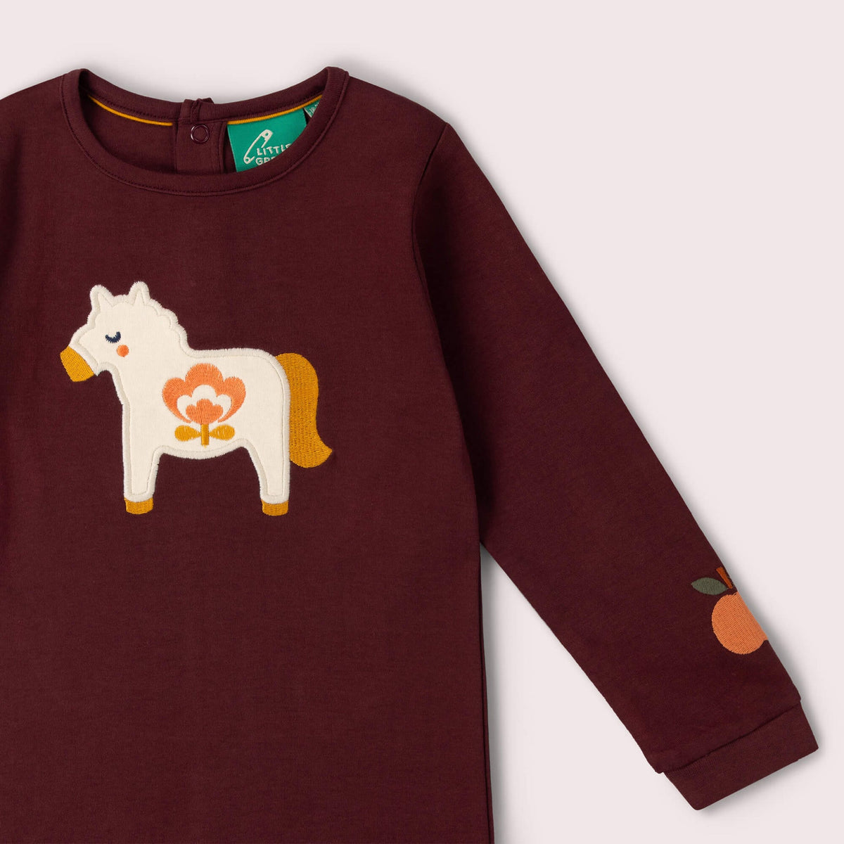 A close-up of a long-sleeve children's top in deep burgundy, featuring a white wooden horse applique with orange and yellow details on the chest and an orange patch on the sleeve.