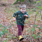 A young boy running outdoors on a leaf-covered path, wearing a dark green long-sleeve sweatshirt with a colorful animal and holiday print, burgundy pants, and yellow rain boots.