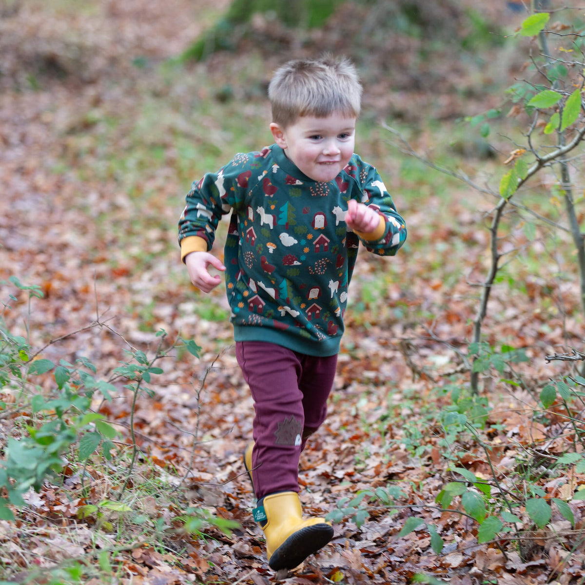 A young boy running outdoors on a leaf-covered path, wearing a dark green long-sleeve sweatshirt with a colorful animal and holiday print, burgundy pants, and yellow rain boots.