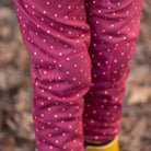 Close-up of a child's legs wearing pink leggings with a light pink polka dot pattern and reinforced knee patches, standing outdoors on a leaf-covered ground.
