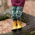 A child's legs wearing green patterned pants and bright yellow rain boots, standing on a mossy tree branch in an outdoor forest setting.