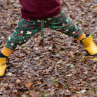A child's legs wearing green patterned pants with autumn-themed prints, yellow rain boots, and black socks, standing on a ground covered in dry brown leaves.