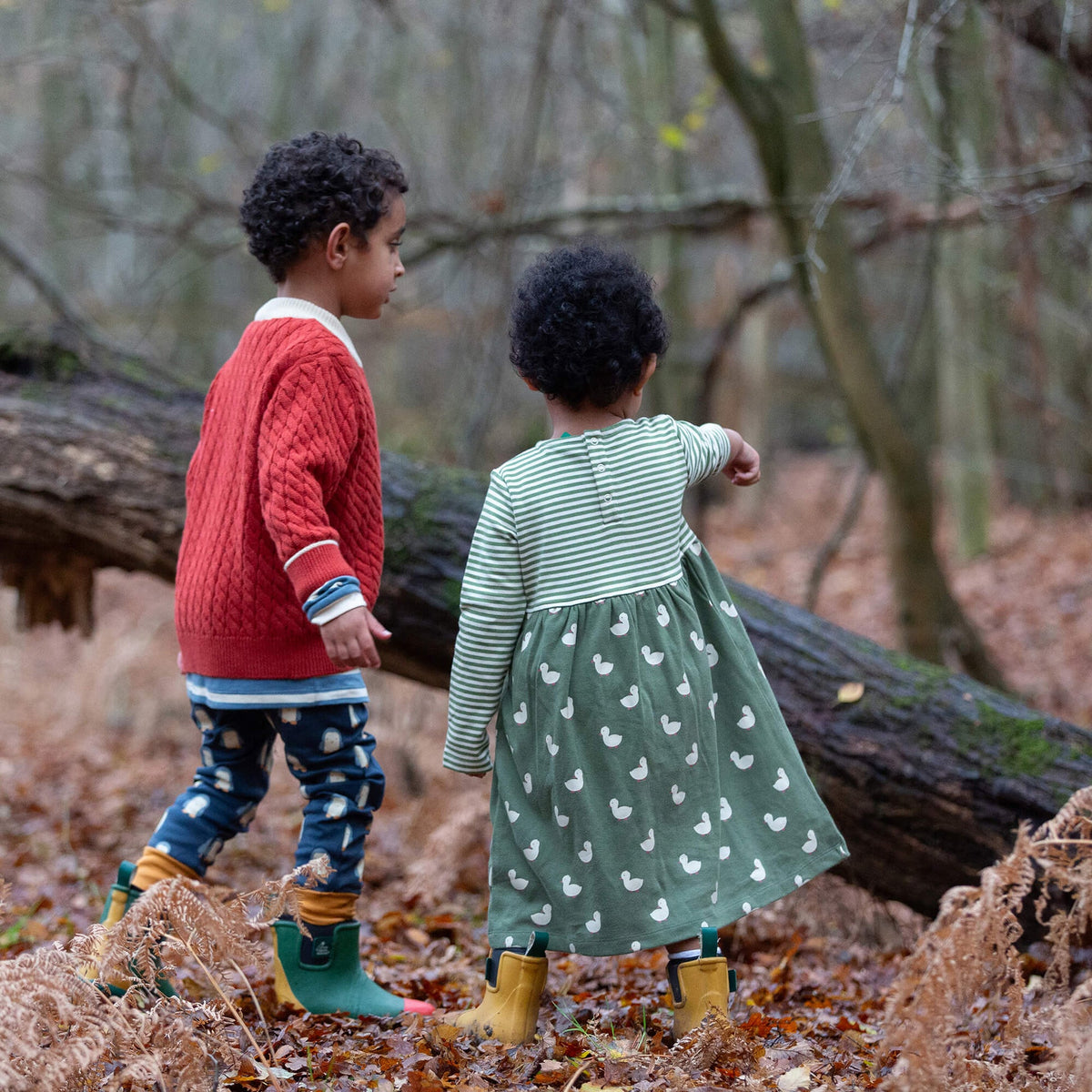 Two young children outdoors in a wooded setting. The child in the foreground is shown from the back, wearing a green and white striped long-sleeve top with a green skirt patterned with white ducks.