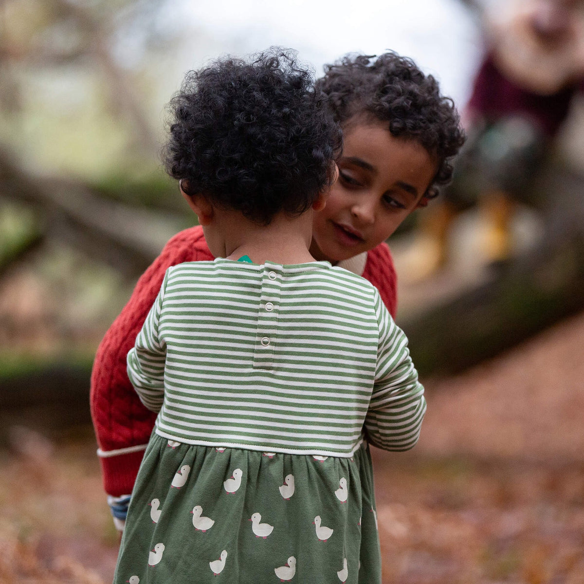Two young children outdoors in a wooded setting. The child in the foreground is shown from the back, wearing a green and white striped long-sleeve top with a green skirt patterned with white ducks.