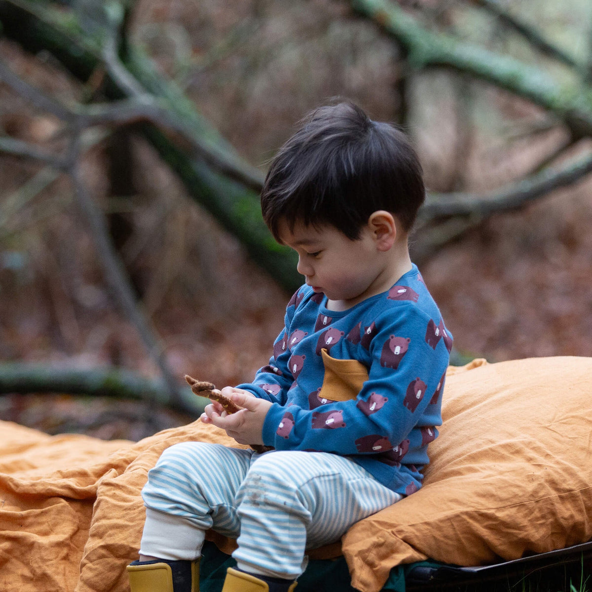 A child sitting and wearing blue long-sleeve shirt with bear pattern and yellow pocket on a blurred background.