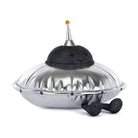 A plush toy shaped like a silver UFO with a black glittery smiling dome top, a metallic silver pointed antenna with a small orange ball, and black glittery feet, photographed on a clean white background.