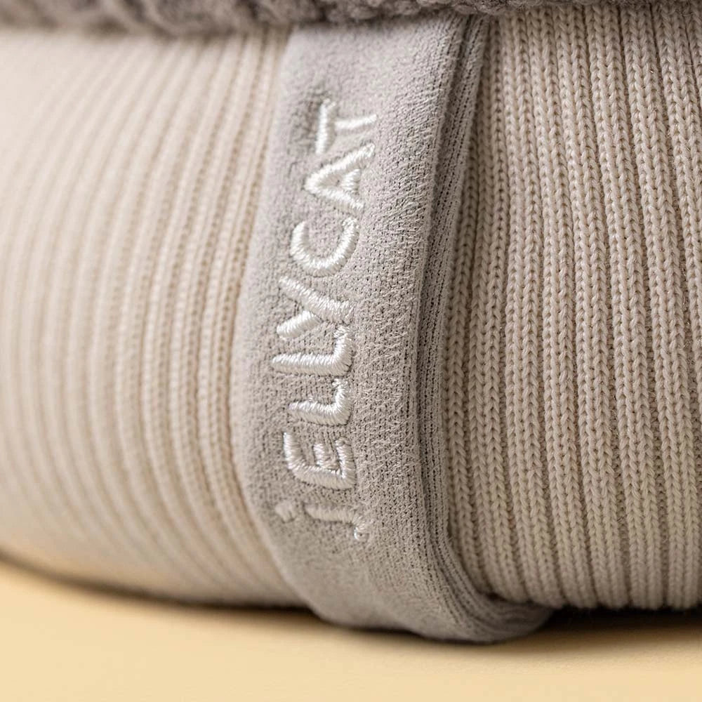 Close-up of a ribbed knit blanket or cushion in a light warm gray-beige color, with the brand name 'JELLYCAT' embroidered vertically on a fabric tab.