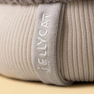 Close-up of a ribbed knit blanket or cushion in a light warm gray-beige color, with the brand name 'JELLYCAT' embroidered vertically on a fabric tab.
