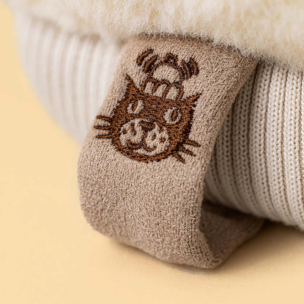 A close-up of a beige knit sock folded over a cream ribbed knit boot or sweater, featuring a brown embroidered cat face with a small hat on the cuff, set against a soft beige background.
