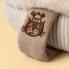 A close-up of a beige knit sock folded over a cream ribbed knit boot or sweater, featuring a brown embroidered cat face with a small hat on the cuff, set against a soft beige background.