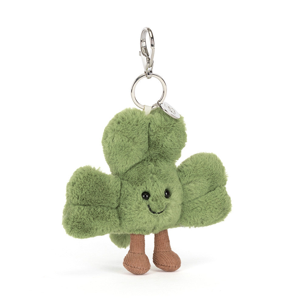 A plush shamrock keychain charm with a smiling face, black bead eyes, and brown fabric feet, attached to a silver metal clasp and ring on a white background.