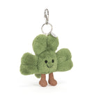 A plush shamrock keychain charm with a smiling face, black bead eyes, and brown fabric feet, attached to a silver metal clasp and ring on a white background.
