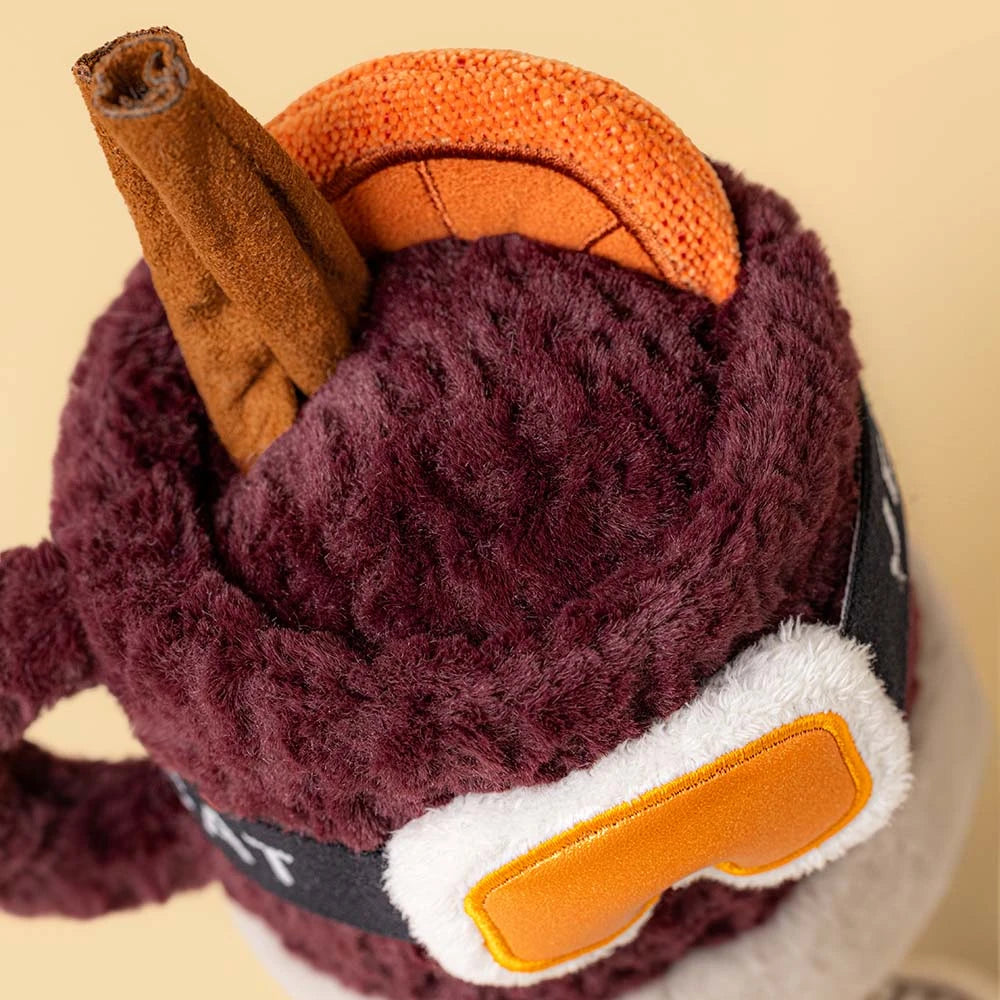 A close-up of a plush toy or stuffed accessory with a dark burgundy fuzzy body, an orange horn-like detail, a brown textured protruding piece, and white-and-orange goggles on a black strap, set against a plain beige background.