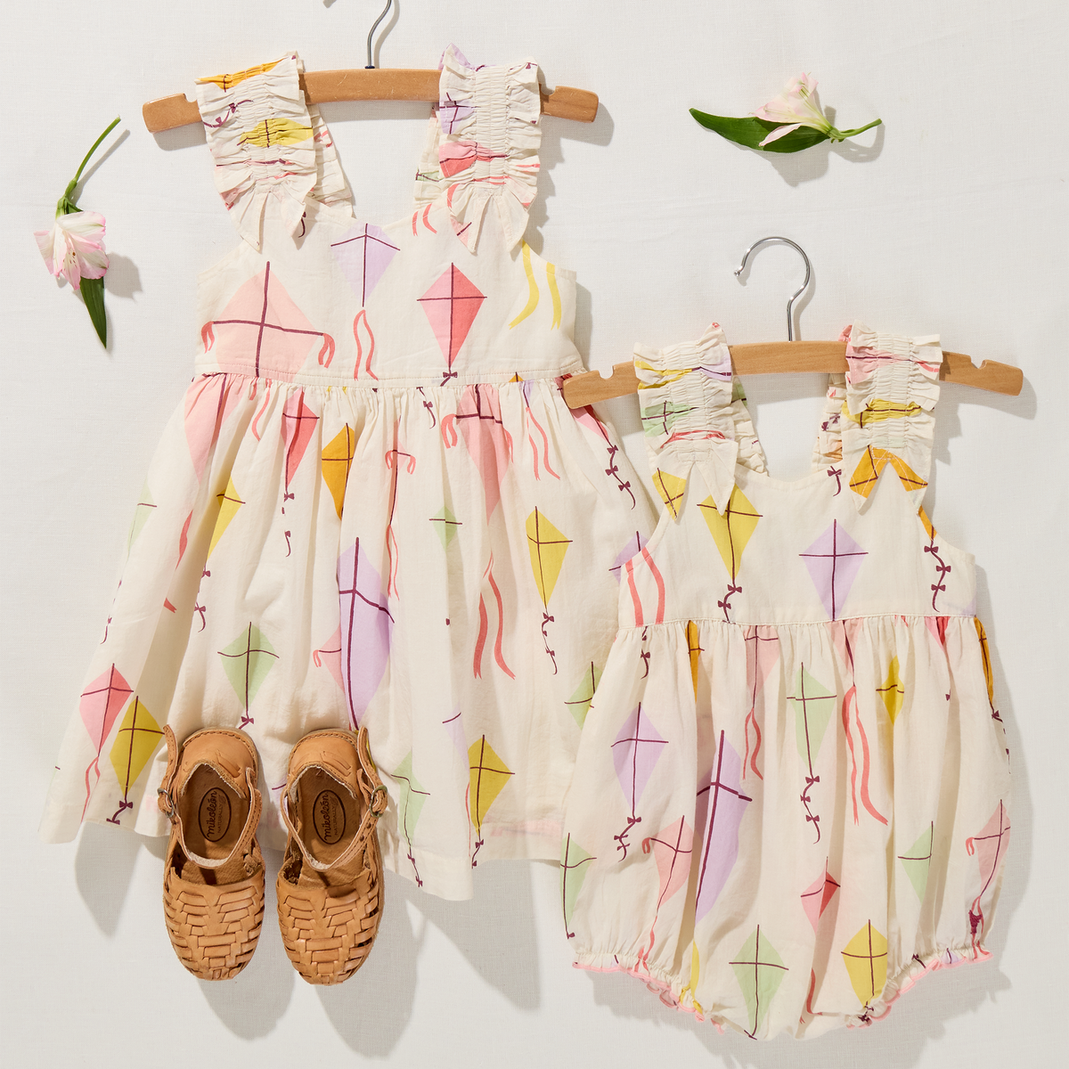 Two children's rompers with kite pattern on hangers, accompanied by a pair of shoes.