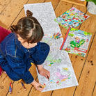 Child coloring a large sheet of paper with coloring books and crayons on a wooden floor.