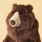 A close-up of a brown plush bear toy with a cream-colored snout and dark eyes, set against a solid pale yellow background.