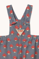Close-up of a children's corduroy overall in a blue-gray color with a red and white mushroom print, featuring a triangular beige patch with the brand name 'TINY' on the chest.
