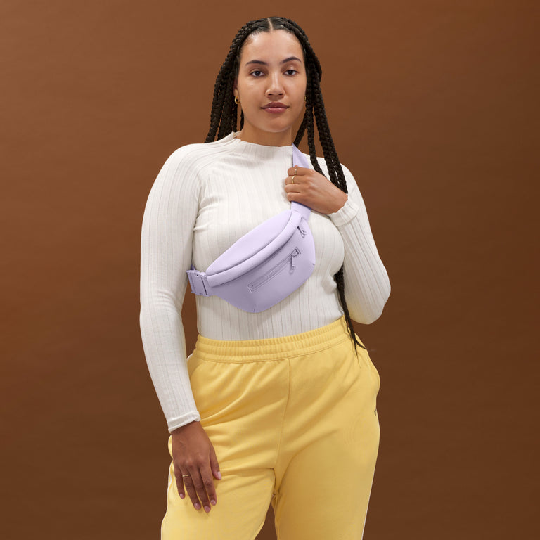 Woman wearing a white sweater and yellow pants with a purple waist bag against a brown background