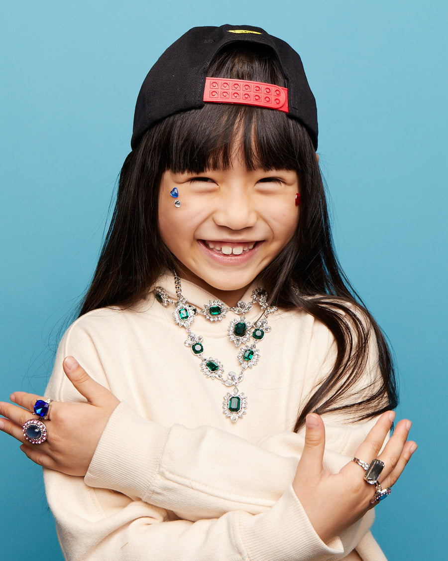 Child wearing a black cap with a red logo, beige sweater, and jewelry against a blue background