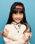 Child wearing a black cap with a red logo, beige sweater, and jewelry against a blue background