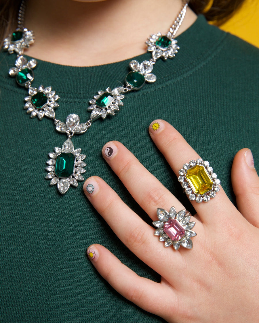 Close-up of a person wearing a green dress with a detailed necklace and a hand with a ring on a yellow background.