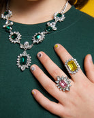 Close-up of a person wearing a green dress with a detailed necklace and a hand with a ring on a yellow background.