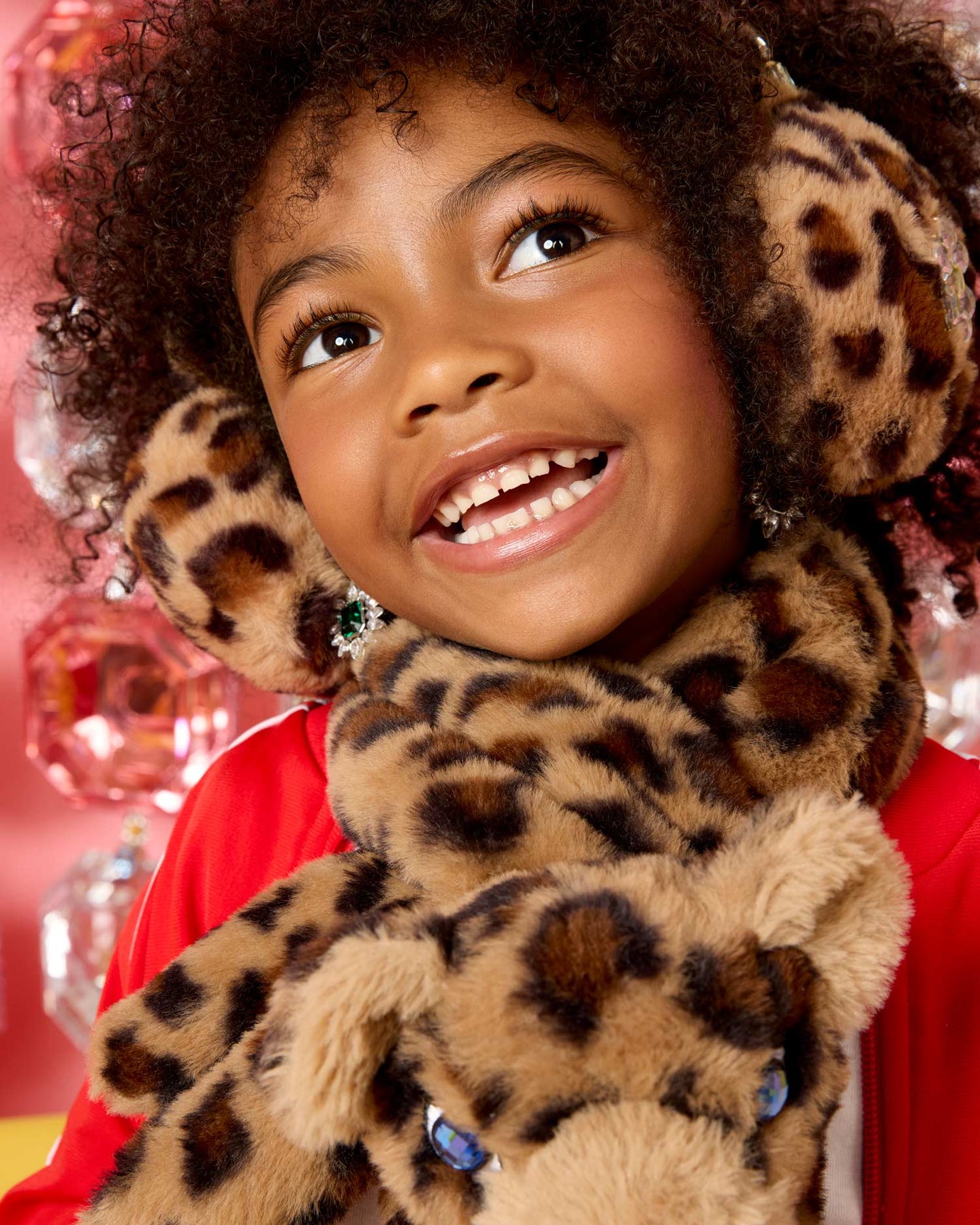 A smiling young girl with curly hair wearing plush leopard-print earmuffs and a matching scarf, with a stuffed leopard toy in front of her face. She is wearing a red top, and the background is a festive pink holiday decor setting.
