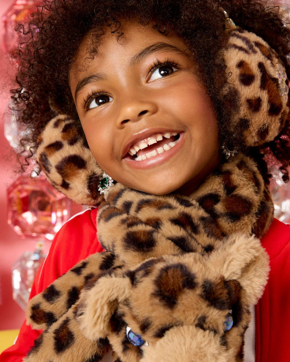 A smiling young girl with curly hair wearing plush leopard-print earmuffs and a matching scarf, with a stuffed leopard toy in front of her face. She is wearing a red top, and the background is a festive pink holiday decor setting.