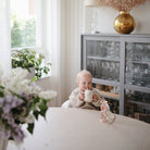 Baby holding a mug in a room with a window, plants, and a glass cabinet.