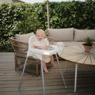 Baby in a high chair on a wooden deck with outdoor furniture and greenery.