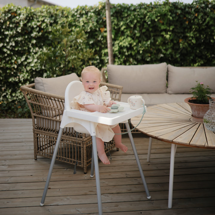 Baby in a high chair on a wooden deck with outdoor furniture and greenery.