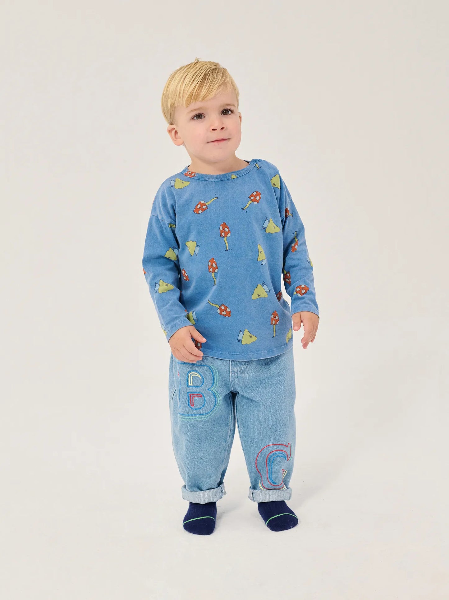 A young child wearing a light blue long-sleeve top with a colorful bug print and matching light blue pants with large embroidered letters, standing against a plain light background.
