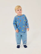 A young child wearing a light blue long-sleeve top with a colorful bug print and matching light blue pants with large embroidered letters, standing against a plain light background.