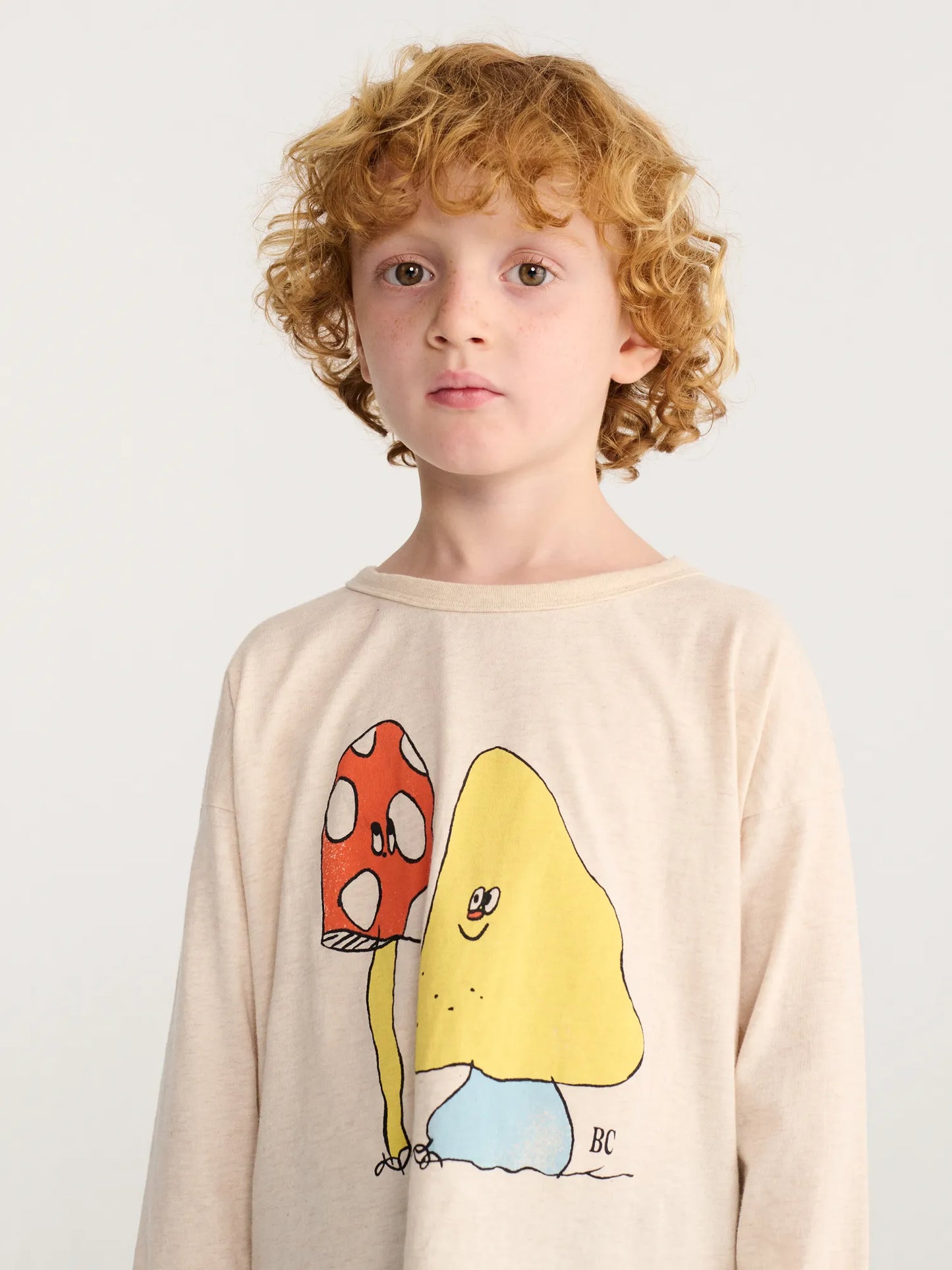 A boy wearing beige long-sleeve crewneck shirt featuring a cartoon graphic of a red-and-white mushroom and a yellow smiling character.
