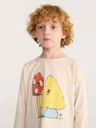 A boy wearing beige long-sleeve crewneck shirt featuring a cartoon graphic of a red-and-white mushroom and a yellow smiling character.
