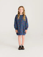 A young girl with long blonde hair stands against a light gray studio background wearing a navy blue crewneck sweatshirt with a red and green cherry pattern, orange and white striped tights, and brown shoes.