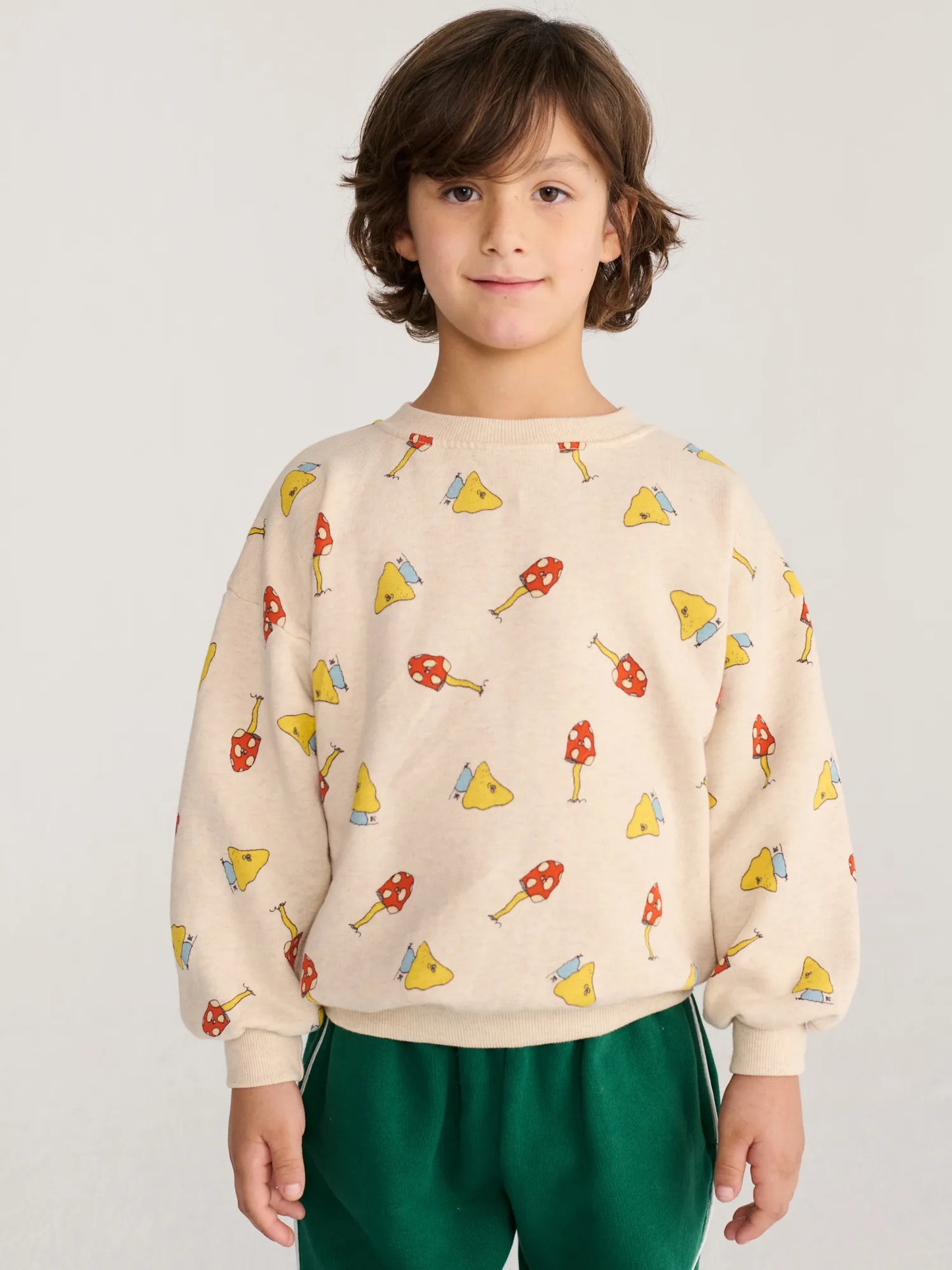 A young boy with brown hair wearing a cream-colored crewneck sweatshirt with a colorful mushroom and cheese slice pattern, paired with green pants, against a light gray background.