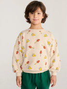 A young boy with brown hair wearing a cream-colored crewneck sweatshirt with a colorful mushroom and cheese slice pattern, paired with green pants, against a light gray background.
