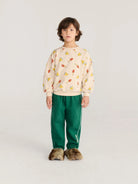 A child model stands against a light gray studio background wearing a cream sweatshirt with a colorful red, yellow, and green pattern, paired with green track pants and brown faux-fur slippers.