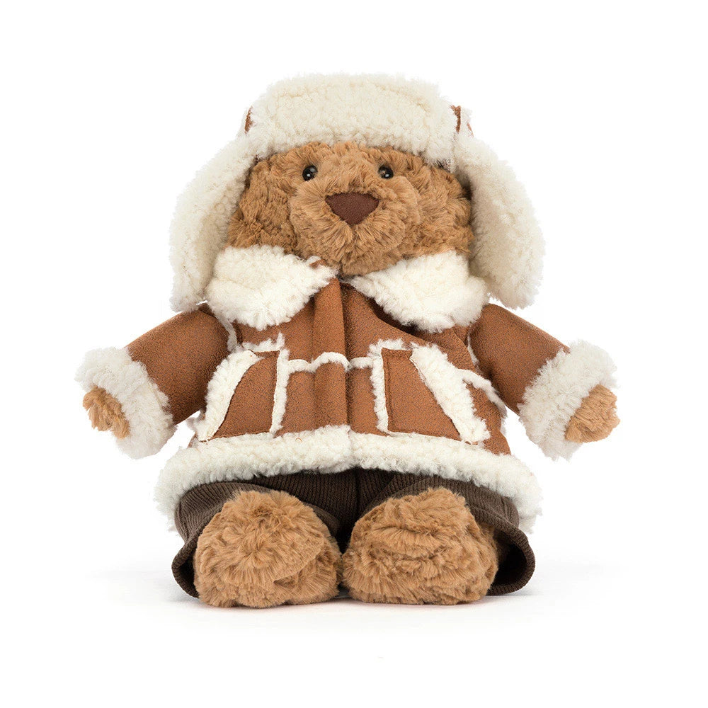 A plush teddy bear wearing a brown faux-leather aviator-style jacket with white faux-fur trim, a matching white aviator hat, and dark brown pants, photographed on a clean white background.