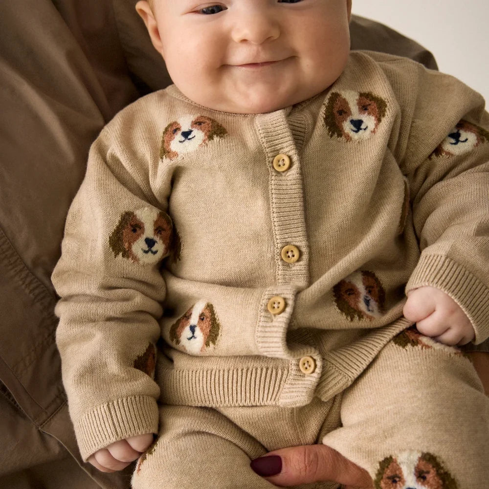 A baby wearing a beige knit cardigan and matching pants with dog face patterns, photographed in a close-up lifestyle setting.