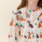 A close-up of a baby dress with a cream-colored base and an all-over print of dachshunds, birthday candles, party hats, balloons, and gift boxes