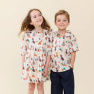 A young girl and boy modeling matching short-sleeve children's shirts and a dress with a colorful animal print on a cream background, set against a solid light beige backdrop.