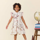 A young girl with curly dark hair stands smiling in a cream-colored dress decorated with a colorful pencil print. She is posed in front of a plain beige background with a stack of books and a globe on a small wooden stand to the right.