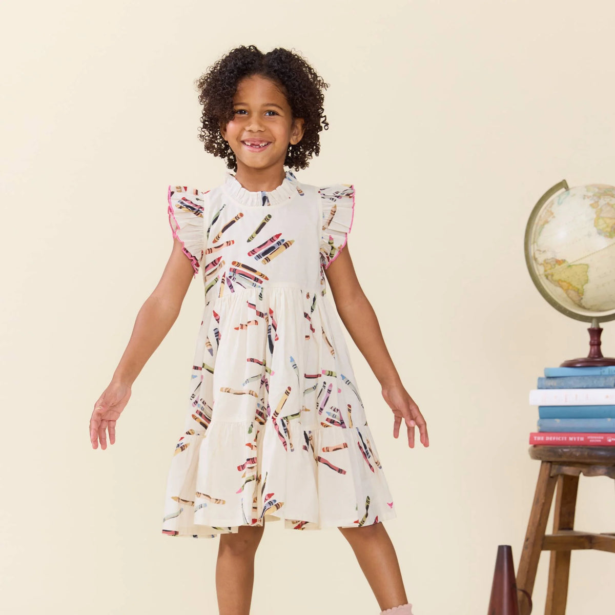 A young girl with curly dark hair stands smiling in a cream-colored dress decorated with a colorful pencil print. She is posed in front of a plain beige background with a stack of books and a globe on a small wooden stand to the right.