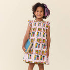 Young girl wearing a colorful dress with a pattern of juice boxes, holding a book against a beige background.