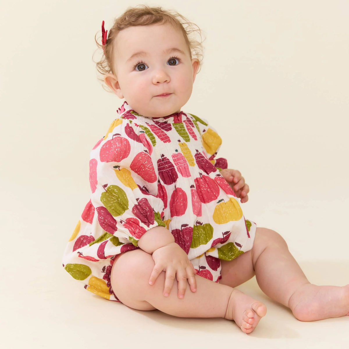 A baby wearing a short-sleeve romper with a colorful fruit print, sitting on a warm cream background.